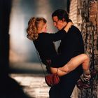 Es gibt keine sichtbaren Texte im Bild. Eine Frau hält einen Mann, die Szene wirkt intim, auf einem Kopfsteinpflasterweg.