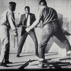 Unten rechts: "HN-L-76". Drei Männer in einer spannungsgeladenen Szene. Einer in Anzug, zwei in Arbeitskleidung mit Ketten.