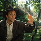 Ein Mann im Abenteueroutfit hält ein kleines Artefakt im Dschungel dicht bei seinem Gesicht. Er schaut gespannt nach oben.