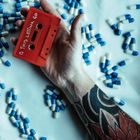 Ein rotes Kassettenband mit der Aufschrift "IS THIS LETTING GO" in einer tätowierten Hand, umgeben von blauen und weißen Kapseln.
