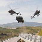 Zwei Helikopter transportieren ein Auto über eine kurvige Autobahn zwischen grünen Weinbergen und Hügeln.