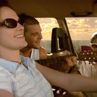 Drei Personen sitzen im Auto, eine spielt Gitarre. Alle tragen Sonnenbrillen, im Hintergrund ein sonniger Himmel.