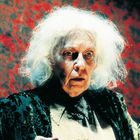 Eine ältere Person mit weißem, wildem Haar, in altmodischer Kleidung, wirkt erschrocken vor einem roten, gemusterten Hintergrund.