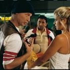 Drei Personen stehen in einer Flughafenhalle. Ein Mann mit Mütze spricht gestikulierend mit einer blonden Frau in weißem Kleid.