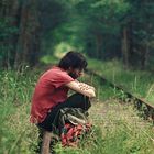 Ein Mensch sitzt auf Bahngleisen in einer grünen, bewaldeten Umgebung und blickt gedankenverloren in die Ferne.