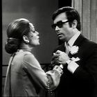 Ein Paar in formeller Kleidung hält sich die Hände. Der Mann trägt eine Sonnenbrille und hat eine Blume im Revers.