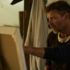 Ein Mann steht konzentriert vor einer Leinwand im Atelier, umgeben von Pflanzen und weichen Tüchern.