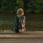 Eine Person sitzt auf einem Holzsteg am Wasser, umgeben von Natur.