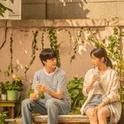 Ein Mann und eine Frau sitzen auf einer Bank im Garten, trinken aus Plastikbechern, umgeben von Pflanzen.