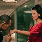 Ein Mann küsst die Hand einer Frau in rotem Kleid. Sie trägt eine Perlenkette und Ohrringe, beide wirken elegant gekleidet.