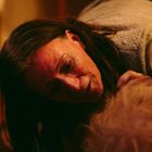 Eine Frau mit langen Haaren blickt sorgenvoll nach unten, nahe bei einer anderen Person in einem warm beleuchteten Raum.