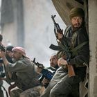 Fünf Männer in Tarnkleidung mit Gewehren kauern in einer Kampfposition in einer städtischen Umgebung.