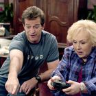 Ein Mann im T-Shirt mit Zeichnungen sitzt neben einer älteren Frau mit Controller, beide wirken konzentriert.