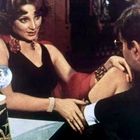 Eine Frau in schwarzem Kleid sitzt an einem Tisch mit einem Mann. Sie hat braune Haare und trägt Schmuck.