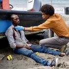 Gelbe Nummernmarkierungen 3 und 1 auf Sand. Ein Mann in blutbeflecktem Hemd lehnt an einem Boot, ein anderer Mann untersucht ihn.