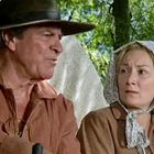 Zwei Personen in historischer Kleidung, ein Mann mit Hut und eine Frau mit Haube, unterhalten sich im Freien.