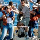 Mehrere Personen auf einem Boot. Eine Person trägt eine Schwimmweste, wird von anderen gestützt. Meer im Hintergrund.