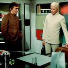 Zwei Männer stehen in einem Raum, einer in braun, der andere in weiß. Ein Schreibtisch vor ihnen, mit Büchern im Hintergrund.