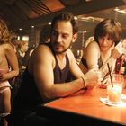 „Red Bull“ Leuchtschrift im Hintergrund. Eine belebte Bar, links eine Frau in Dessous, rechts ein Paar mit Getränken.