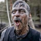 Ein Mann mit öligem Gesichtsausdruck schreit. Er trägt einen Anzug, im Hintergrund sind Bäume und Metallstrukturen.