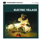 Text: "ELECTRIC VILLAGE". Ein Mann sitzt hinter drei beleuchteten Lampen, trägt einen Turban und sieht ernst aus.