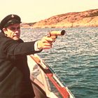 Ein Mann in Uniform und Mütze steht auf einem Boot und hält eine Pistole. Im Hintergrund sind Wasser und eine felsige Küste sichtbar.