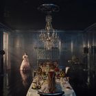 In einem prunkvollen Raum mit dunklem Boden steht ein gedeckter Tisch unter einem Kronleuchter. Eine Frau in Rosakleid betritt.