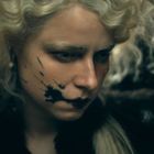 Eine Person mit hellblonden, lockigen Haaren und schwarzem Fleck-Design auf der Wange, in dunkler Kleidung.