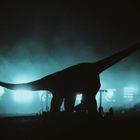 Ein dunkler Dinosaurier-Schatten vor hell erleuchtetem Hintergrund, Menschen im Vordergrund, mystische Atmosphäre.