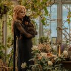 Eine Frau mit roten Haaren hält eine schwarze Katze in einem üppigen Gewächshaus voller Pflanzen und Blumen.