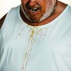 Ein Mann in einem weißen Shirt hat flüssige Spritzer darauf. Sein Gesicht und Bart sind nass und er wirkt angespannt.