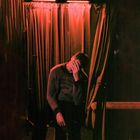 Person sitzt in rotem Licht vor Vorhängen, Gesicht verdeckt. Text auf Pullover: "Acne Studios STOCKHOLM".