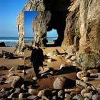 Ich kann keine Texte im Bild erkennen. Zu sehen ist ein Strand mit großen Felsen und ein Torbogen aus Stein. Eine Person geht am Strand entlang.