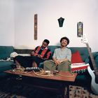 Zwei lachende Männer sitzen auf einem blauen Sofa vor einem Tisch mit Kabeln und Instrumenten; eine gemütliche Wohnzimmeratmosphäre.