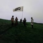 Eine Gruppe Menschen in winterlicher Kleidung steht auf einer Wiese. Sie tragen Instrumente; einer hält eine Fahne mit zwei schwarzen Wellenlinien.