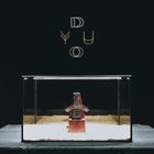 Text: "DUYO" in großen Buchstaben. Ein Metronom steht in einem Glasbehälter auf hellem Sand. Dunkler Hintergrund.