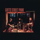 "Gotts Street Park" und "Somewhere Between" stehen oben. Drei Musiker spielen in einem gemütlichen Raum, umgeben von Dunkelheit.