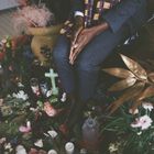 Eine Person sitzt mit gefalteten Händen, umgeben von Blumen und Kerzen; ein grünes Kreuz ist sichtbar.