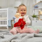 Ein lächelndes Baby sitzt auf einer Decke, hält einen Teddybär und ein rotes Tuch. Im Hintergrund ist ein Kinderbett.