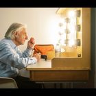 Eine ältere Person sitzt an einem beleuchteten Schminktisch und schaut in den Spiegel.