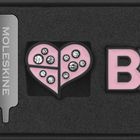 Ein graviertes Metallobjekt mit "MOLESKINE", ein pinkes herzförmiges Symbol mit Steinen und "BP" aus rosa Buchstaben.
