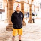 Ein Mann mit pinkem Irokesenschnitt und Bart, trägt schwarzen Hoodie mit kleinem Emblem, gelbe Shorts, tätowiertes Bein.