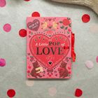 „A Little POP of LOVE“. Pop-up Buch mit Herz, zwei Bären und Rosen, auf grauem Hintergrund mit roten Konfetti.