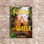 "FOLLOW THE WATER" von Ellen Cochrane, Teenager in Wasser mit Stock im Amazonas; grüner Wald und Flugzeug darüber.