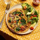 Ein Teller mit einem bunten Omelett, umgeben von Salat, Avocado und Tomaten auf einem gelben Tischset. Links ein Besteck.