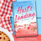 Buchtitel "Hart's Landing" von Melanie Harlow, ein rot-weißes Tuch, ein Stück Kuchen auf blauem Holz.