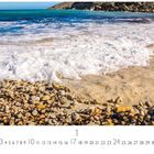 "Am Strand", darunter ein Monatskalender. Steiniger Strand mit schäumenden Wellen, blauem Meer und Felsen im Hintergrund.