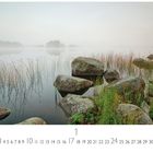 Morgennebel am See. Eine ruhige Landschaft mit Steinen und Gras am Ufer eines nebligen Sees.