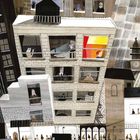 Illustration einer Stadtansicht mit geneigten Häusern, Menschen in Fenstern und auf der Straße, Autos und einem Uhrturm.