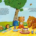 Zwei Bären auf einer Wiese, der eine sitzt, der andere liegt. Ein Picknick mit Kuchen, Tassen und Tellern. Ein Rucksack mit einem Vogel darauf.